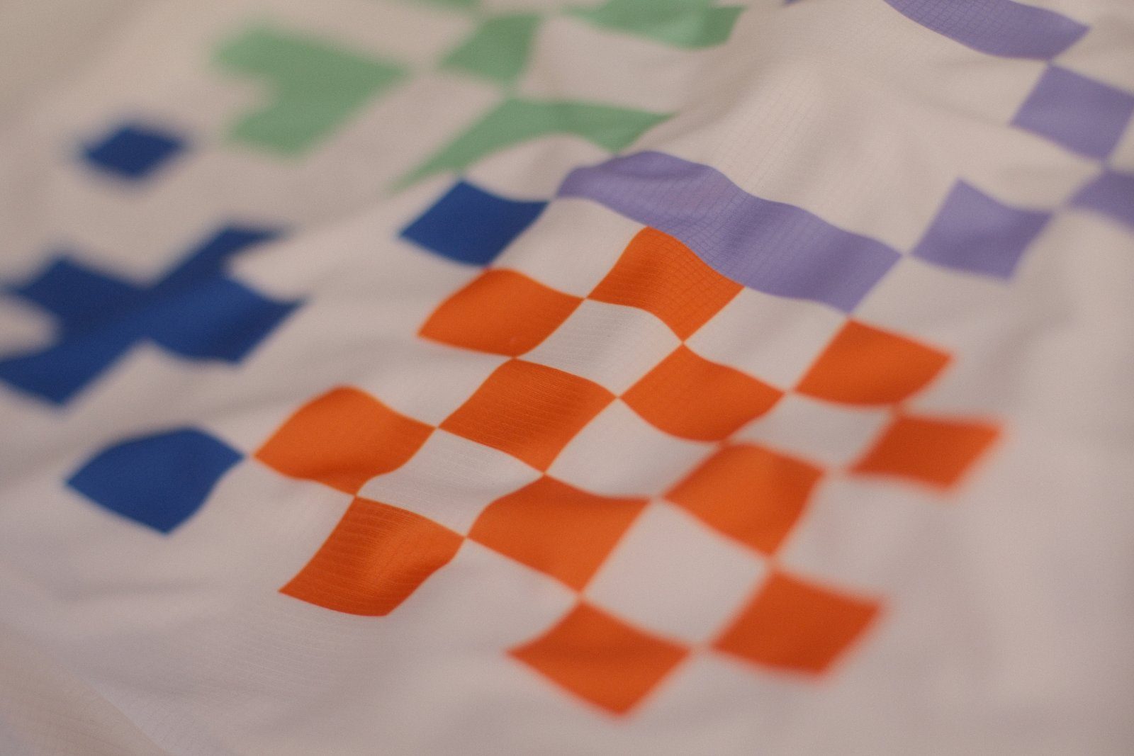 Detailaufnahme eines Sublimationsdrucks auf weißem Funktionsshirt: orange, blaue, grüne und violette Raster-Kacheln, die Farbe sitzt direkt im Gewebe