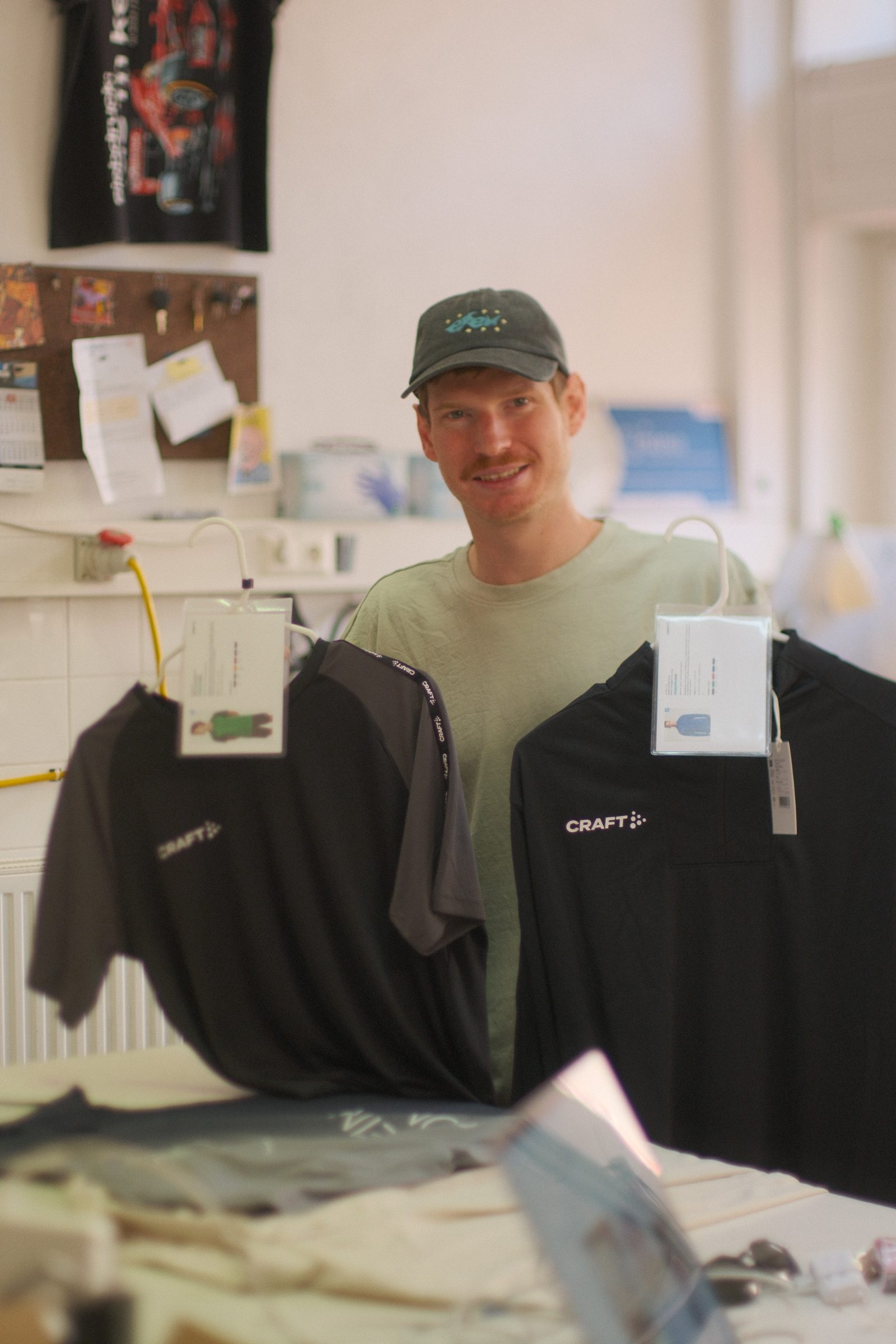 Markus hält zwei schwarze Craft-Funktionsshirts mit Etiketten in der Werkstatt von Aus'm Keller
