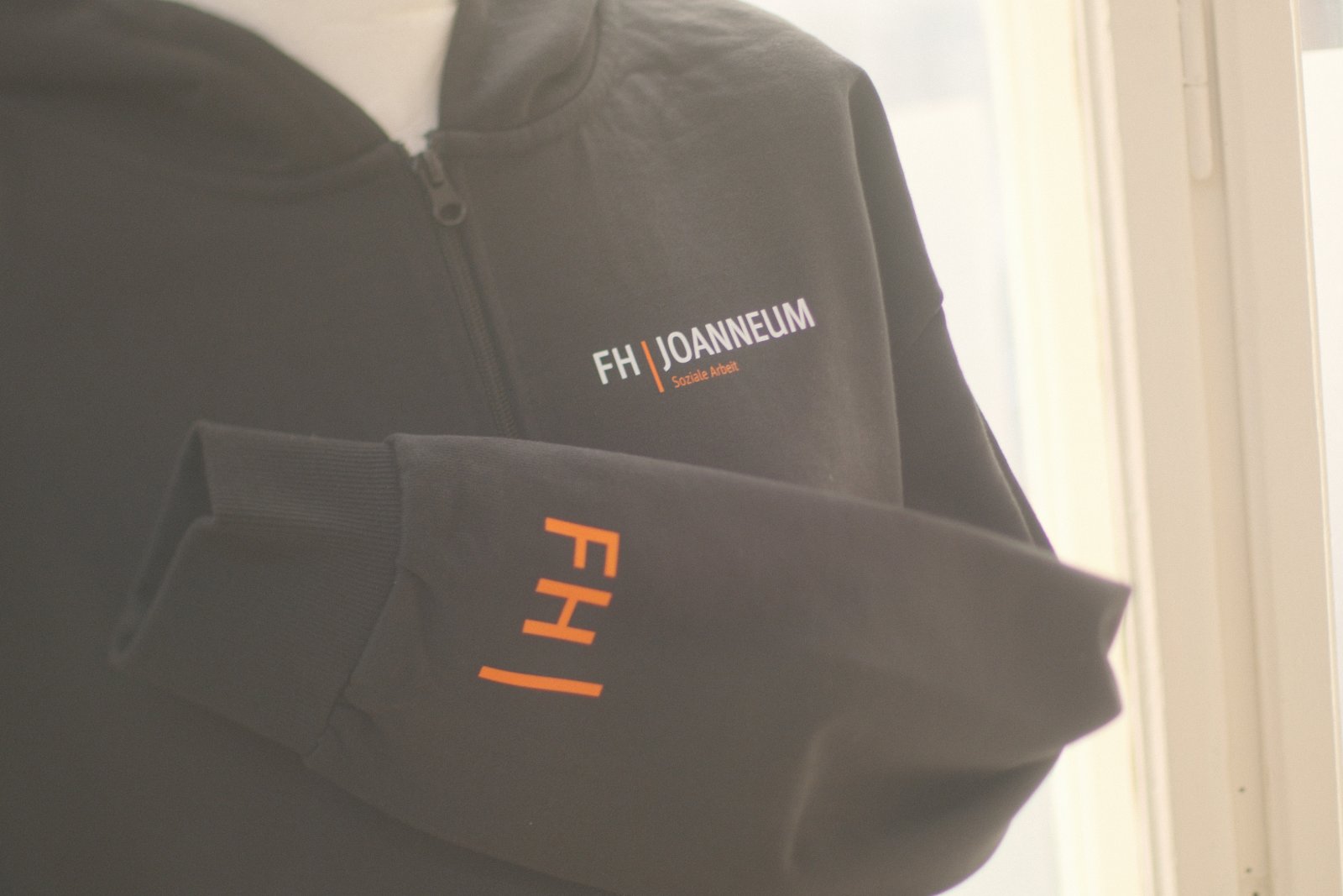 Schwarze Zipperjacken der FH Joanneum Soziale Arbeit mit weißem Brustlogo und orangem FH-Akzent am Ärmel