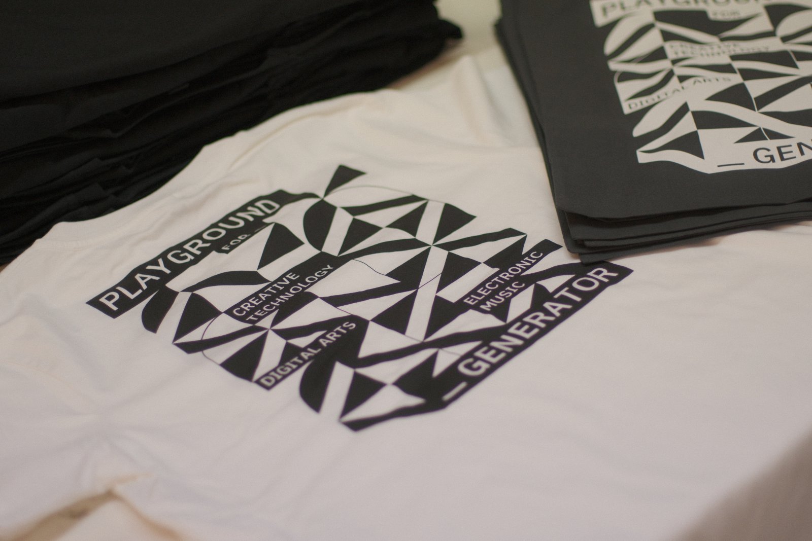 Stapel der Generate26-Festival-Shirts in zwei Farbvarianten — schwarz und naturweiß, bedruckt im wasserbasierten Siebdruck mit Living Ink Algae-Ink