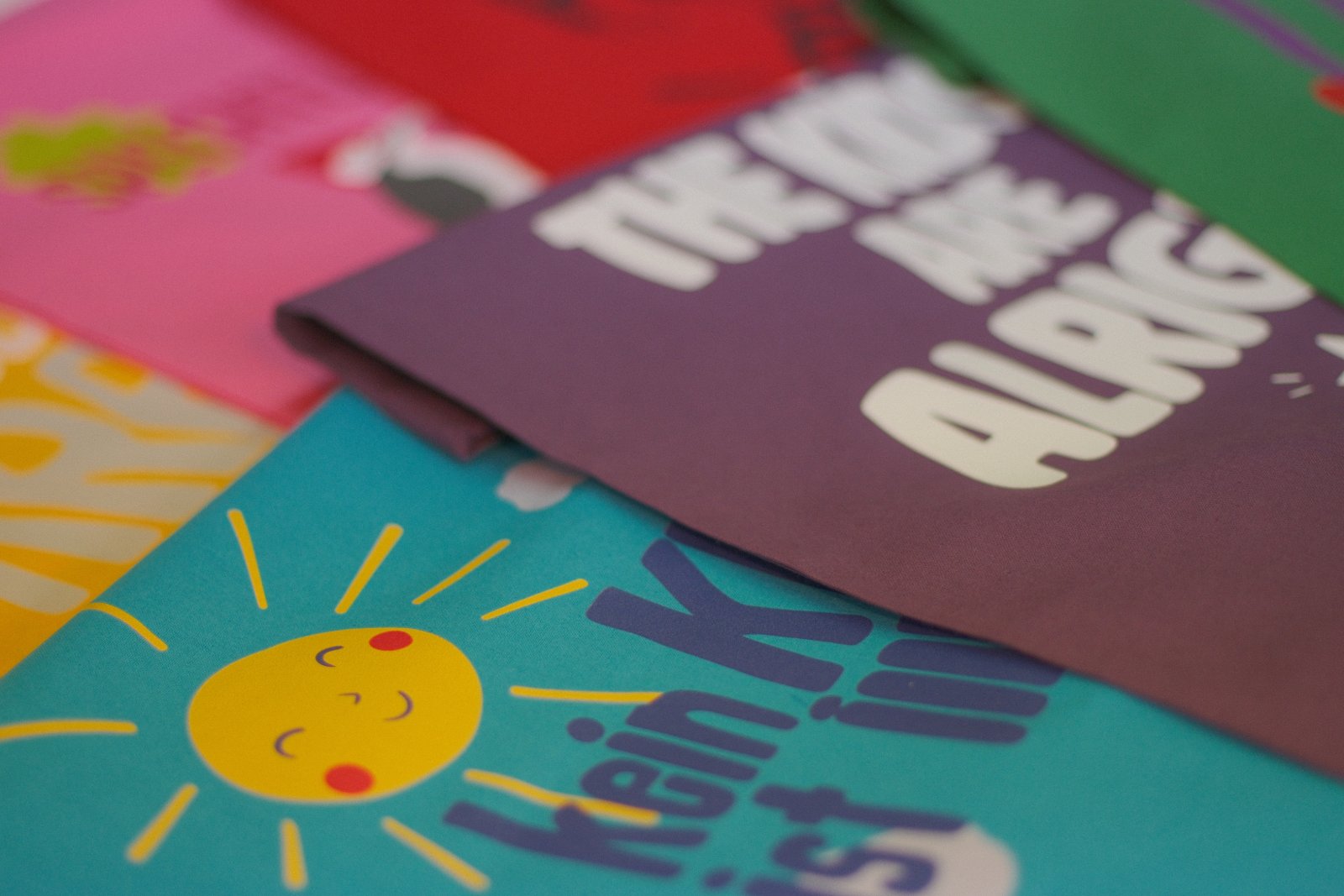 Stapel der bedruckten Kampagnen-T-Shirts in Hellblau, Rot, Pink, Naturweiß, Gelb und Grün — Siebdruck mit Sonnen-Motiv und Slogan 'Kein Kind ist illegal'