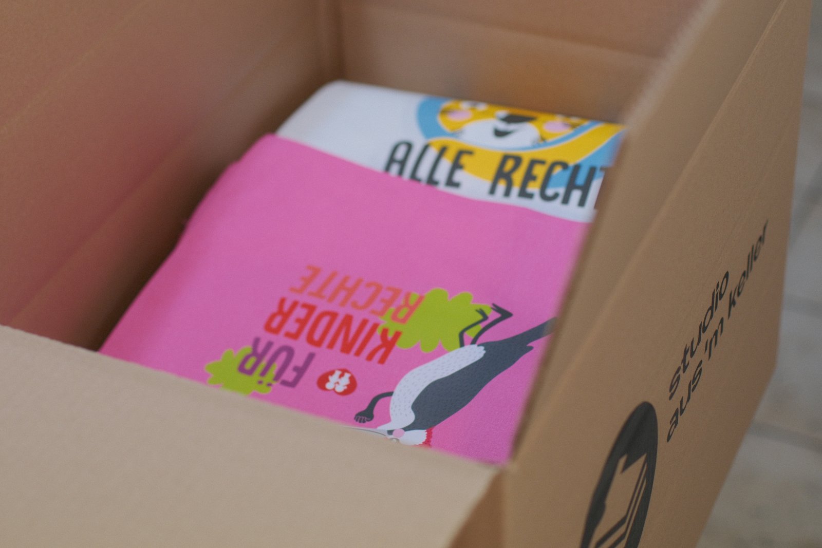 Fertig verpackte Auflage im Aus'm Keller-Versandkarton — Pinkes DTF-Shirt mit Motiv 'Für Kinderrechte' liegt obenauf