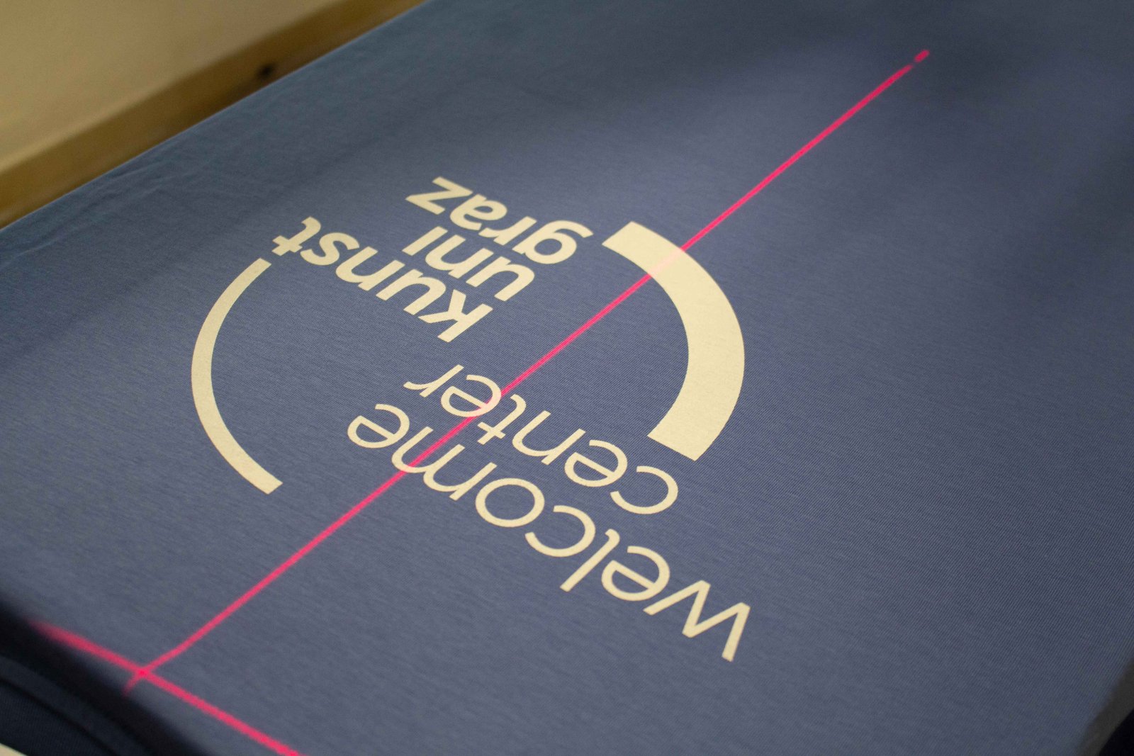Blaues Kunstuni-Graz-Shirt auf der Druckpresse mit Laser-Ausrichtung, Front-Motiv „welcome center