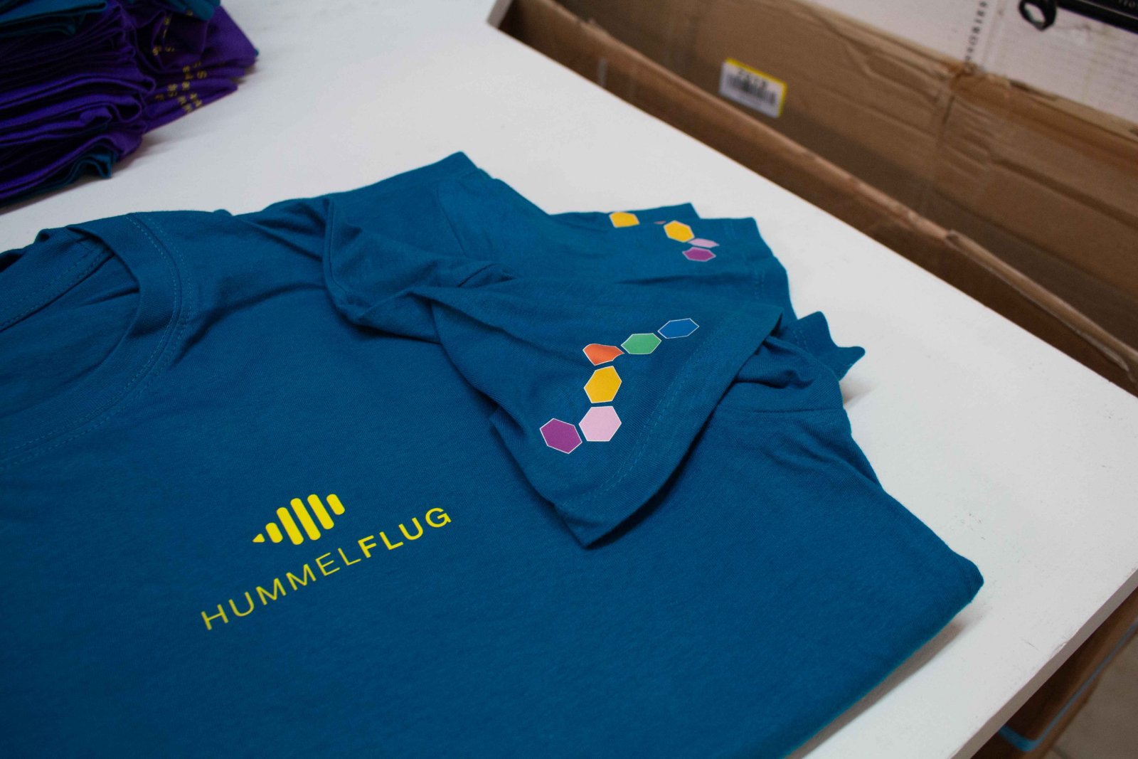Blaues Hummelflug-Shirt der Raiffeisen-Landesbank Steiermark mit gelbem Logo im Siebdruck und bunten Hexagon-Akzenten am Ärmel