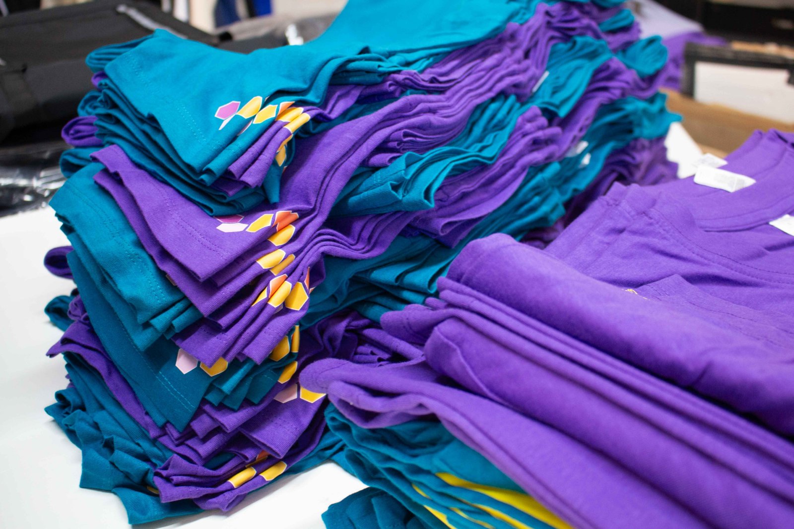 Stapel der Hummelflug-Shirts in Petrol-Blau und Lila mit bunten Hexagon-Akzenten an Schulter und Ärmel