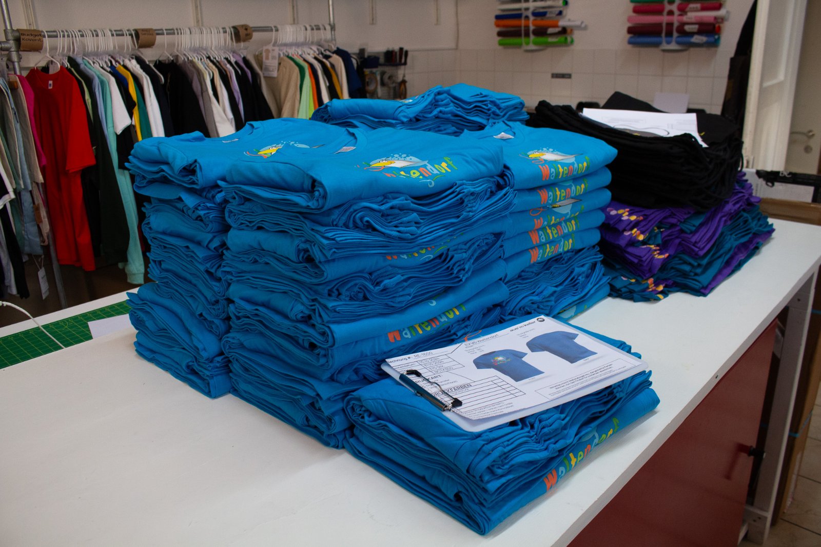 Großer Stapel blauer Kindershirts der VS Waltendorf mit Pelikan-Maskottchen im Digital-Siebdruck-Transfer auf dem Konfektionstisch im Studio