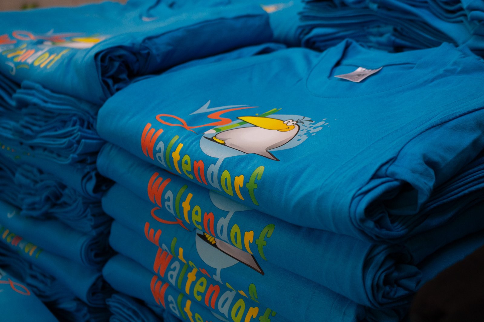 Detailaufnahme der bedruckten Kindershirts — Pelikan-Maskottchen mit Bleistift und bunter Waltendorf-Schriftzug auf Petrol-Blau, gestochen scharfer DTF-Druck